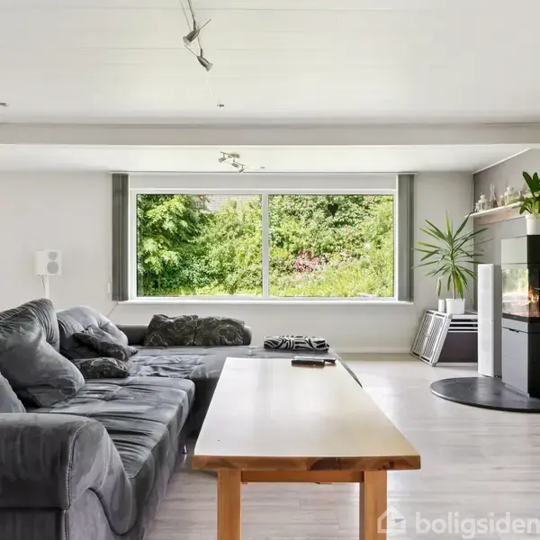 Grå sofa står ved siden af et træbord i en rummelig stue. Rummet har store vinduer med udsigt til frodige buske. Fladskærms-tv er monteret på væggen, og pejsen er tændt.