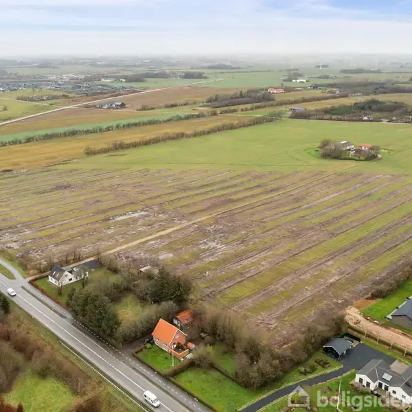 En stor, bar mark strækker sig i mønstrede rækker, omgivet af spredte huse og veje. Landskabet viser et landligt område med åbne marker og få træer under en overskyet himmel.