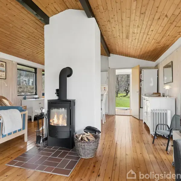 Brændeovn i hjørne på trægulv, omgiven af en hyggelig stue med blå sofa og spisebord i baggrunden. Trævægge og loft tilfører en varm atmosfære. Åben dør viser grøn have.