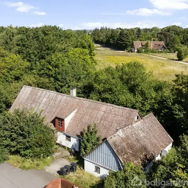 Et ældre hus med skråt tag omgivet af tæt skov og buske. En smal asfaltvej løber ved siden af huset. I baggrunden ses åbne marker og flere huse.