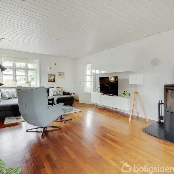 Stue med sort brændeovn til højre, grå stol og sofa til venstre, trælaminatgulv, hvidmalede vægge og loft. Der er et TV monteret på væggen og store vinduer bagved.