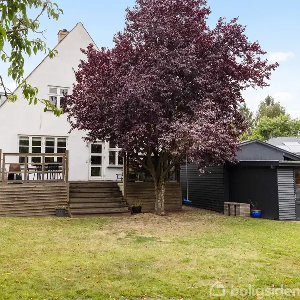 Et hvidt hus med trappe og terrasse actioner ved en stor have. Et lilla træ står tæt på huset, og en grå garage er ved siden af.