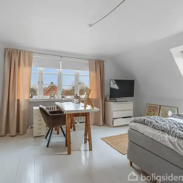 En seng står i et lyst rum med skråvægge. Et bord med stole er placeret ved vinduet, mens en reol og et tv er i nærheden. Gardiner hænger ved vinduet.