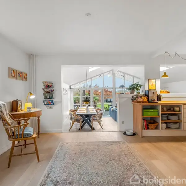 En rød murstenshus står stille mellem to lignende huse. Huset har en trekantet gavl og flere vinduer. Foran ses en hæk, og der er en flisebelagt gangsti til højre.