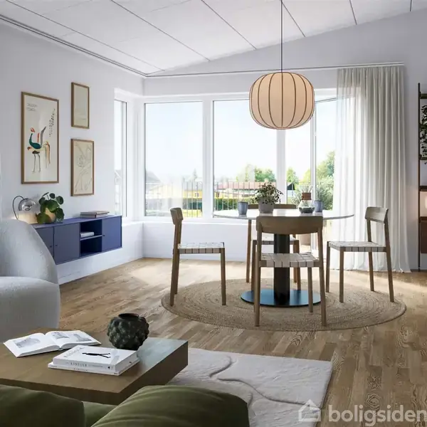 Spisebord med fire stole står på tæppe i velbelyst stue. Store vinduer omkranser rummet, der også har en moderne lænestol, reol og vægbilleder.