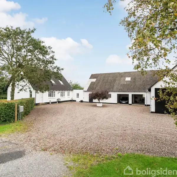 Hvidt hus med mørkt stråtag står stille ved en grusbelagt gårdsplads, omgivet af grønne marker og træer under en blå himmel med spredte skyer. Ingen synlig tekst.