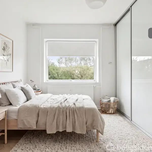 Et soveværelse med en redt dobbeltseng ved siden af et natbord med en plante. Store skydedøre til garderobe, et tæppe på gulvet, og et vindue med udsigt til greenery.