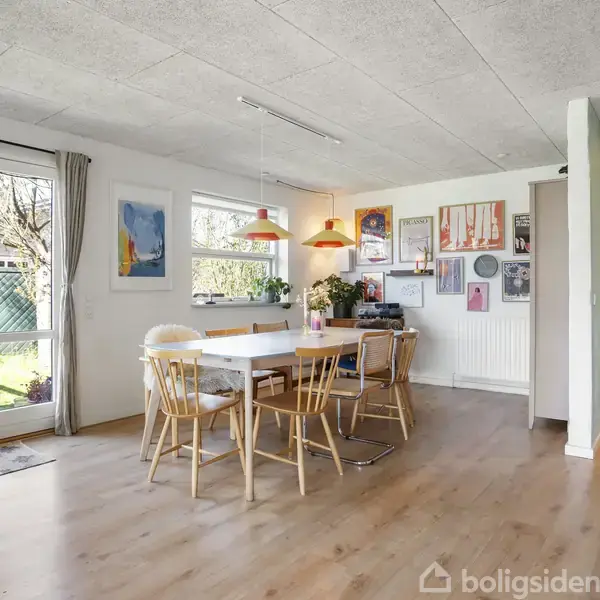 Et lyst spisestueområde indeholder et bord med seks stole. Væggene er dekoreret med kunst, og der er store vinduer med udsigt til en have. Der hænger to orange lamper over bordet.