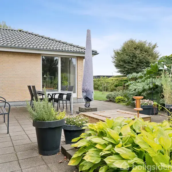 Terrasse med fliser har havemøbler og parasol. Omgivet af grønne planter og buske, ligger ved gulstenshus med havedør, og udsigt til velplejet have med træer og græsplæne.