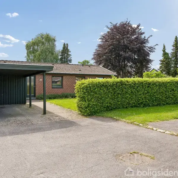 En rødstensvilla med carport står ved en trimmet hæk i et grønt boligområde med grusvej og store træer i baggrunden. Pæn have og solskinsvejr.
