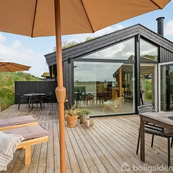 Et moderne sommerhus i sort træ står på en træterrasse. Terrassen har liggestole, et bord med stole og parasoller, omgivet af græs og en klar blå himmel.