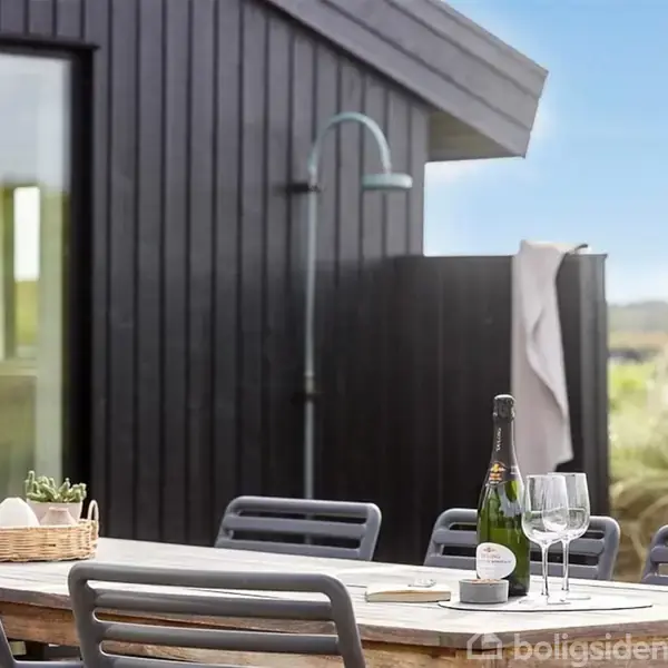 Et bord er dækket med en vinflaske og glas, omgivet af moderne stole. I baggrunden ses en sort træbygning med en udendørs bruser, alt sammen i en naturskøn udendørs omgivelser.