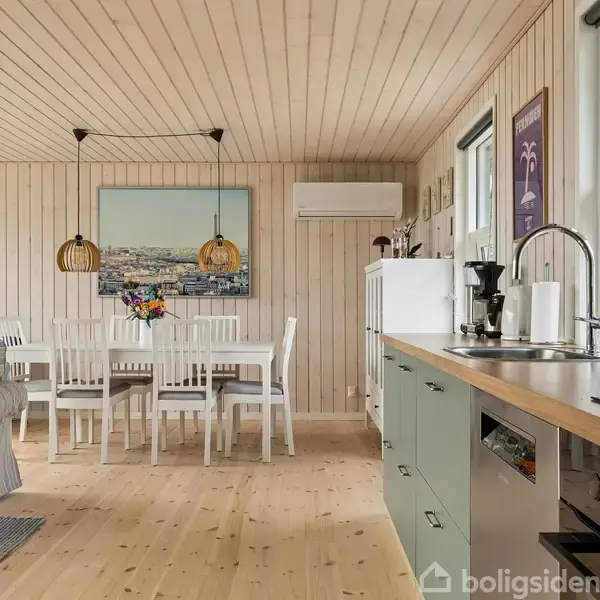 Køkkenalrum med lyse trævægge og -loft. Hvidt spisebord med seks stole står ved siden af en sofa. Vægdekorationer og planter pynter rummet, og der kommer lys ind fra vinduerne.