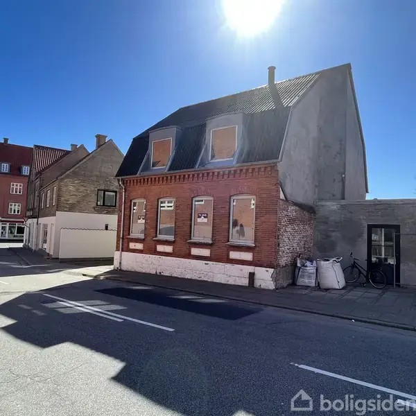 Et rødt murstenshus med tilmurede vinduer står på et gadehjørne under en blå himmel. Ved siden af ses en cykel og nogle hvide sække på fortovet.