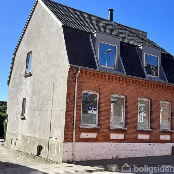 En ældre murstensbygning med sort tag ligger i en by. Vinduerne har "Til Salg"-skilte. Den ene side er hvid, og husets nummer er 1a. Solen skinner klart.