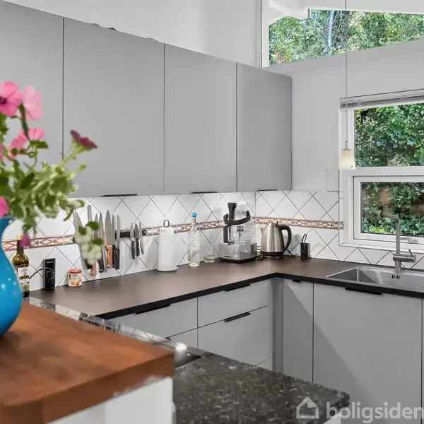 Køkken med grå skabe og sort bordplade. Køkkenredskaber er placeret ved væggen. En blå vase med blomster står i forgrunden. Et vindue viser grønne træer udenfor.