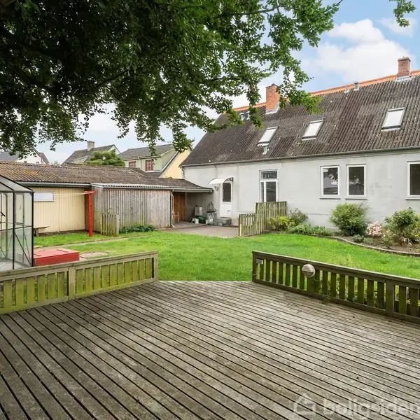 En hvid murstensbygning med skråt tag står bag en træterrasse. I haven er et drivhus og græs arealer omgivet af træer og buske.