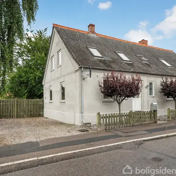 Et hvidt hus med rødt tag står på en stille gade omgivet af et lavt træhegn. Der er små træer foran huset og en grusindkørsel til venstre.