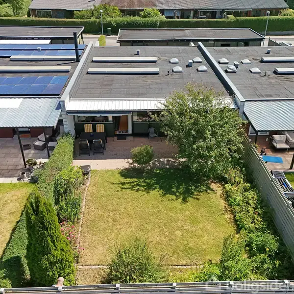 Et rækkehus med sort tag har en baghave med græsplæne, træer og buske. Terrassen har havemøbler, omgivet af andre lignende huse med solpaneler på taget i et villakvarter.
