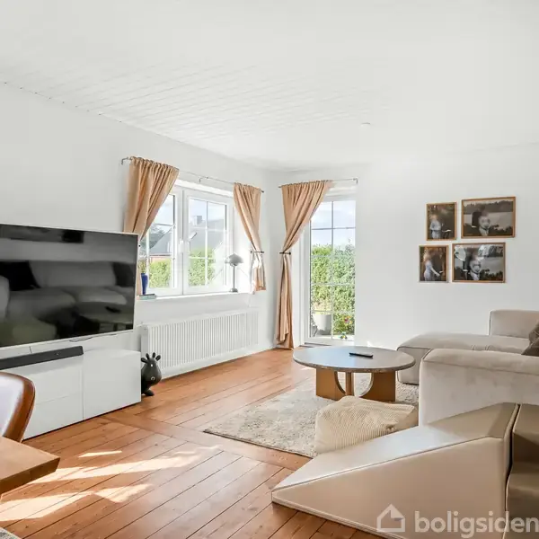Fladskærms-tv står på hvidt skab, mens en sofa og sofabord er placeret i lys stue med træparketgulv og store vinduer med gardiner.