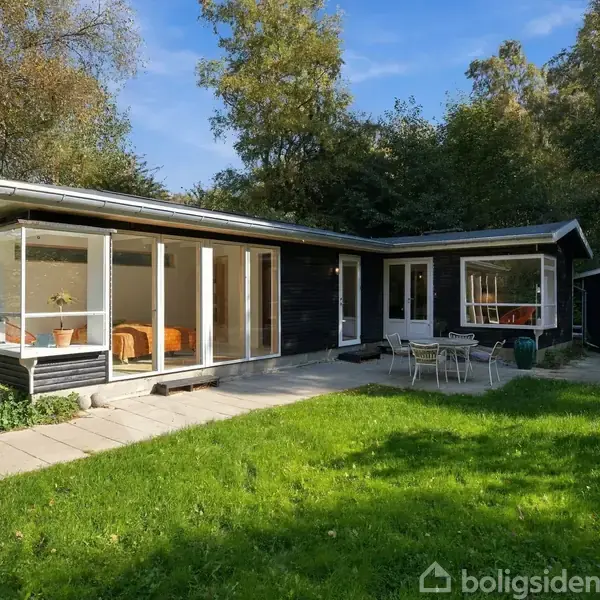 Et sortmalet sommerhus med store vinduer står stille i en solrig have, omgivet af grønne træer. Foran huset er en terrassesten med et hvidt bord- og stolesæt.