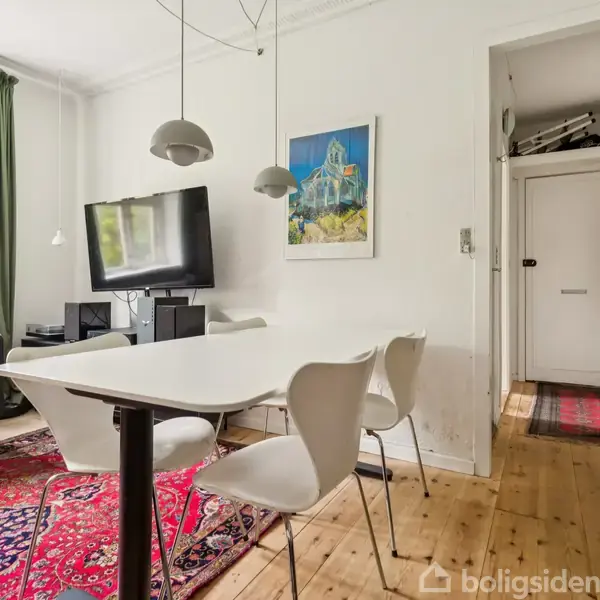 Spisebord med fire stole står på et trægulv. En sofa og et tv ses ved siden af, hængende lamper over bordet. Rød tæppe dækker gulvet i en lys stue.