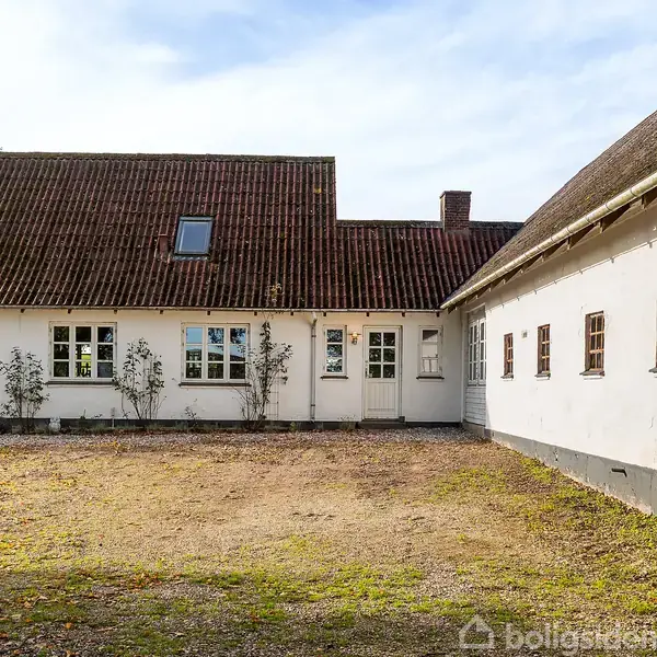 En hvid bygning med brun tagsten står stille i en gårdsplads omgivet af grus. Træer er synlige i baggrunden under en klar himmel.