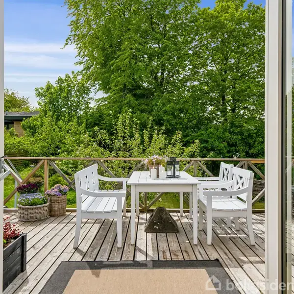 Hvide udendørsmøbler står på en træterrasse. Et bord og stole er centreret. Grønne planter omkranser terrassen, som har udsigt til frodige træer. En døråbning ses i forgrunden.