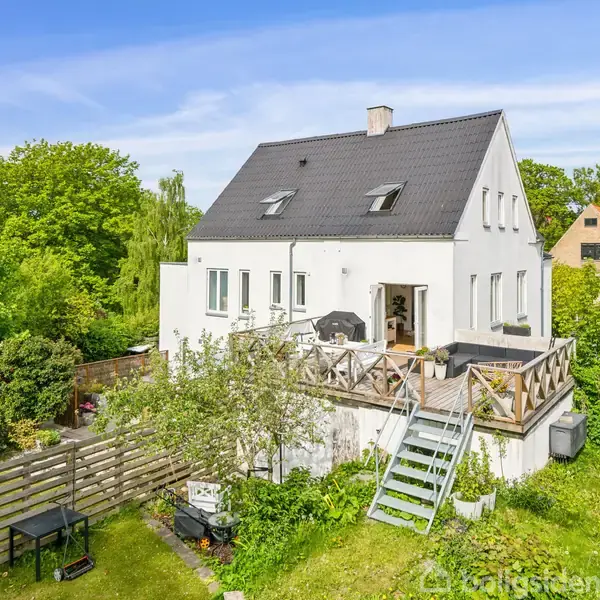 Et hvidt hus med sort tag står i en frodig have. En trappe leder op til en terrasse med havemøbler. Omkring er der grønne træer og buske.