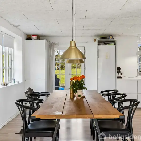 Træbord med seks sorte stole omkring, dekoreret med blomster i vase. Ophængt lampe over bordet i en lys spisestue med vinduer og køkken i baggrunden.