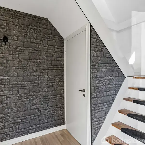 Dør placeret i en entré med murstensvæg og trætrappe ved siden af. Trappen har trin med mørke tæpper. Veggen har en sort trearmet lampe. En plante står ved trappeafsatsen.