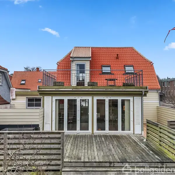 Toetagers hus med rødt tag og stor altan øverst, omgivet af træhegn. Der er en træterrasse foran huset, og en flagstang står til højre i billedet.