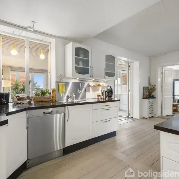 Hvidt køkken med stålopvaskemaskine og kaffemaskine ved siden af vindue. Lys træbordplade og trægulv. Åbning til stue med maleri. Rummets dekorative elementer skaber en lys, hjemlig atmosfære.