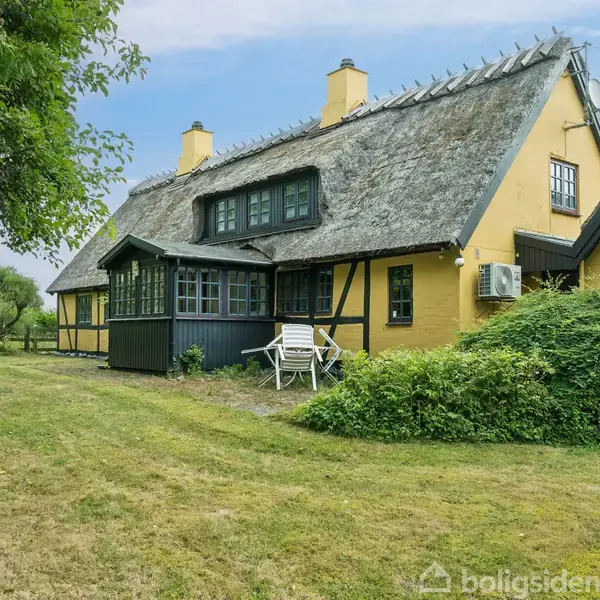Et gult bindingsværkshus med stråtag står omgivet af græs og træer. Der er en hvid stol og et bord foran huset. Sollys skaber en rolig atmosfære.