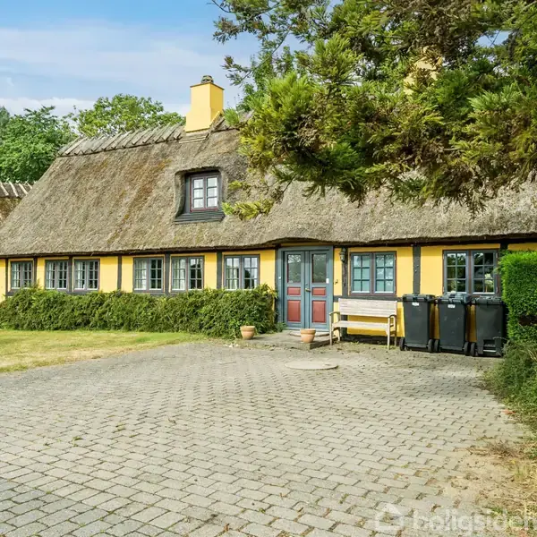Gult bindingsværkshus med stråtag står stille. Foran huset er en stenbelagt gårdsplads, og til højre ses nogle skraldespande. I baggrunden er grønne træer og buske synlige.