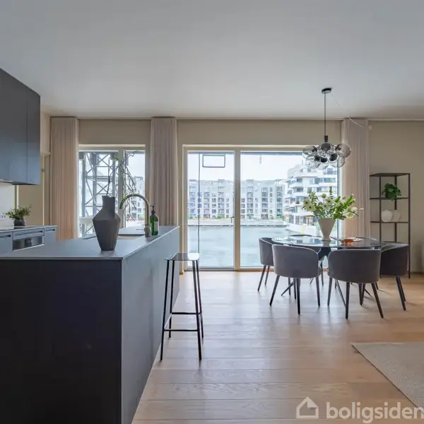 Køkkenø med sorte skabe omgivet af spisebord og stole. Store vinduer viser moderne bygninger udenfor. Lamper hænger over bordet, og sofagruppe ses i baggrunden.