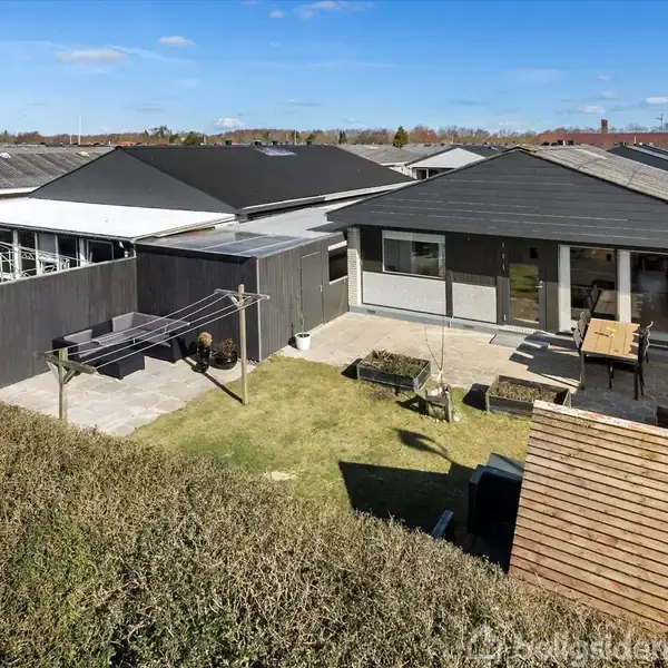 En enkeltstående bolig med fladt tag og store vinduer, står stille i en velplejet have med terrasse og siddepladser. Omgivet af hække og ligger i et roligt boligområde.