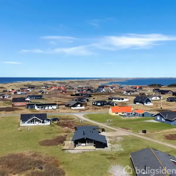 Sommerhuse spredt over et grønt område ved kysten, flere huse har mørke tage. Den klare blå himmel understreger den åbne og fredelige atmosfære nær havet.