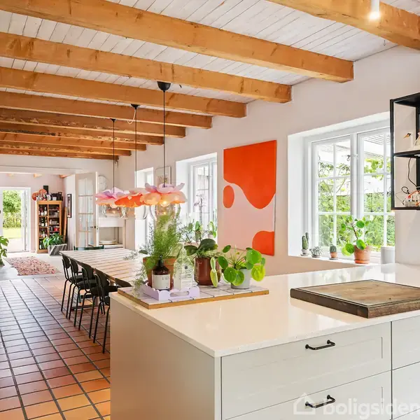 Et køkken-alrum med hvidt køkkenbord og grønne planter står centralt. Loftet har synlige træbjælker. En orange og hvid kunstplakat hænger på væggen. Bord og stole ses længst væk.