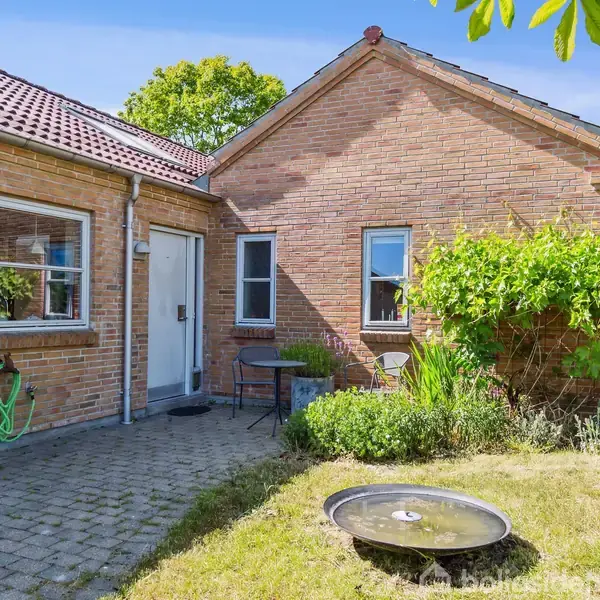 En murstensbygning med en flisebelagt terrasse, hvor der står to stole og et bord. En grøn haveslange hænger på væggen. Omgivet af græsplæne og grønne buske i solskinsvejr.