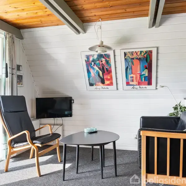 To stole og et bord står i en stue med træloft og hvidmalede vægge. To farverige plakater hænger på væggen, og en lille tv-skærm ses ved siden af.