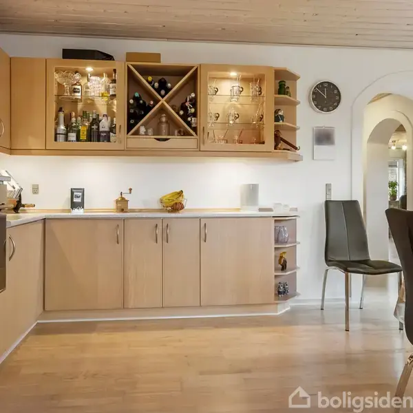 Køkken med lyse træskabe, indbygget ovn og vinklasse opbevaring. Skabene er åbne og indeholder glas og flasker. Spisebord og stole står ved siden af, med lysindfald fra vinduer.