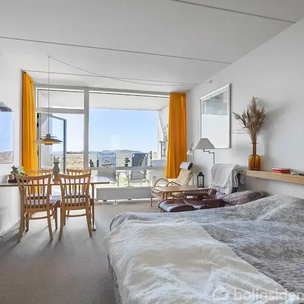 En seng med en grå dyne står i et lyst værelse. Ved siden af er et spisebord med stole placeret foran en stor vindue med gule gardiner, der viser udsigt udenfor. En fladskærm hænger på væggen.