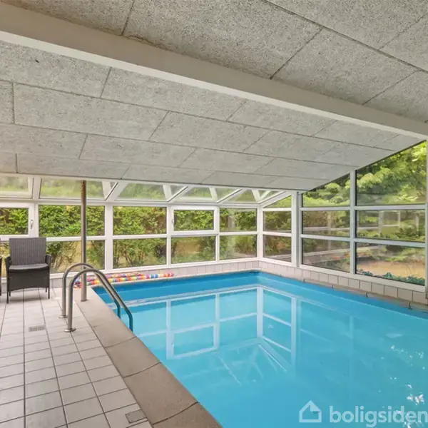 Indendørs swimmingpool med klart vand omgivet af store vinduer. Ved siden af poolen står to sorte stole og et lille bord med en flaske og glas. Udenfor ses træer.