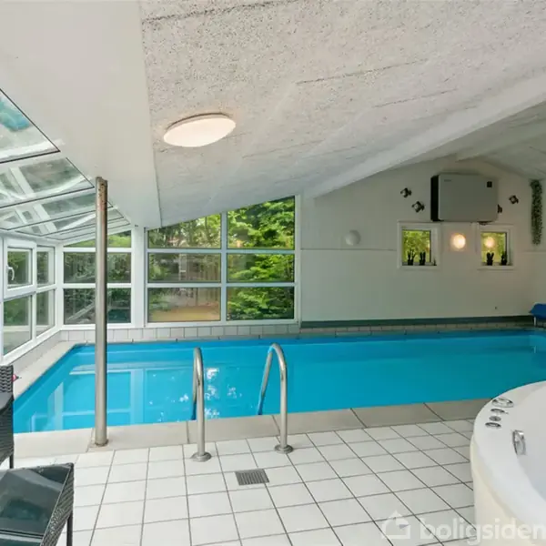 Indendørs swimmingpool fyldt med vand er omgivet af vinduer, med en jacuzzi i nærheden, placeret på flisegulv. Korridortype rum med udsigt til en grøn have udenfor.