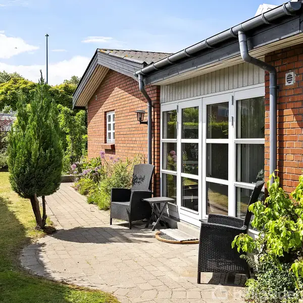Murstenshus med store glasdøre fører ud til terrasse med møbler. Haven er frodig med buske og et træ. Klar blå himmel og solrigt vejr.