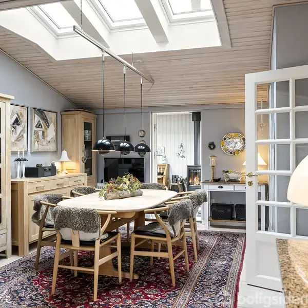 Rundt spisebord med træstole dækket af fåreskind, placeret under sorte pendellamper i et lyst rum. Omgivet af vitrineskabe, tæppe og glasdør, med ovenlys i loftet.