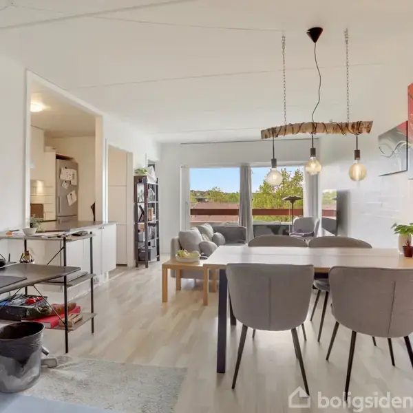 Spisebord med grå stole står i en lys stue. Over bordet hænger tre lamper. I baggrunden ses en sofa foran store vinduer med udsigt til grønt område.