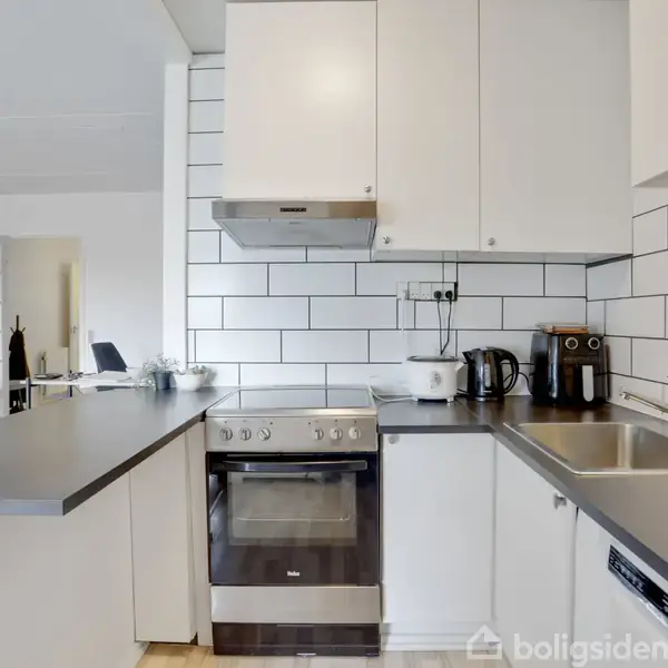 Et moderne køkken har hvidt flisebelagt backsplash, hvide skabe, komfur, vask, og sorte bordplader. Elektriske apparater som elkedel og kaffemaskine står på bordet. Gulvet er lyst trægulv.