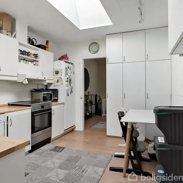 Køkken med hvidt interiør, herunder skabe, en ovn og et køleskab. Et spisebord med stole står ved siden af. Ovenlys oplyser rummet med trægulv og tæppe.